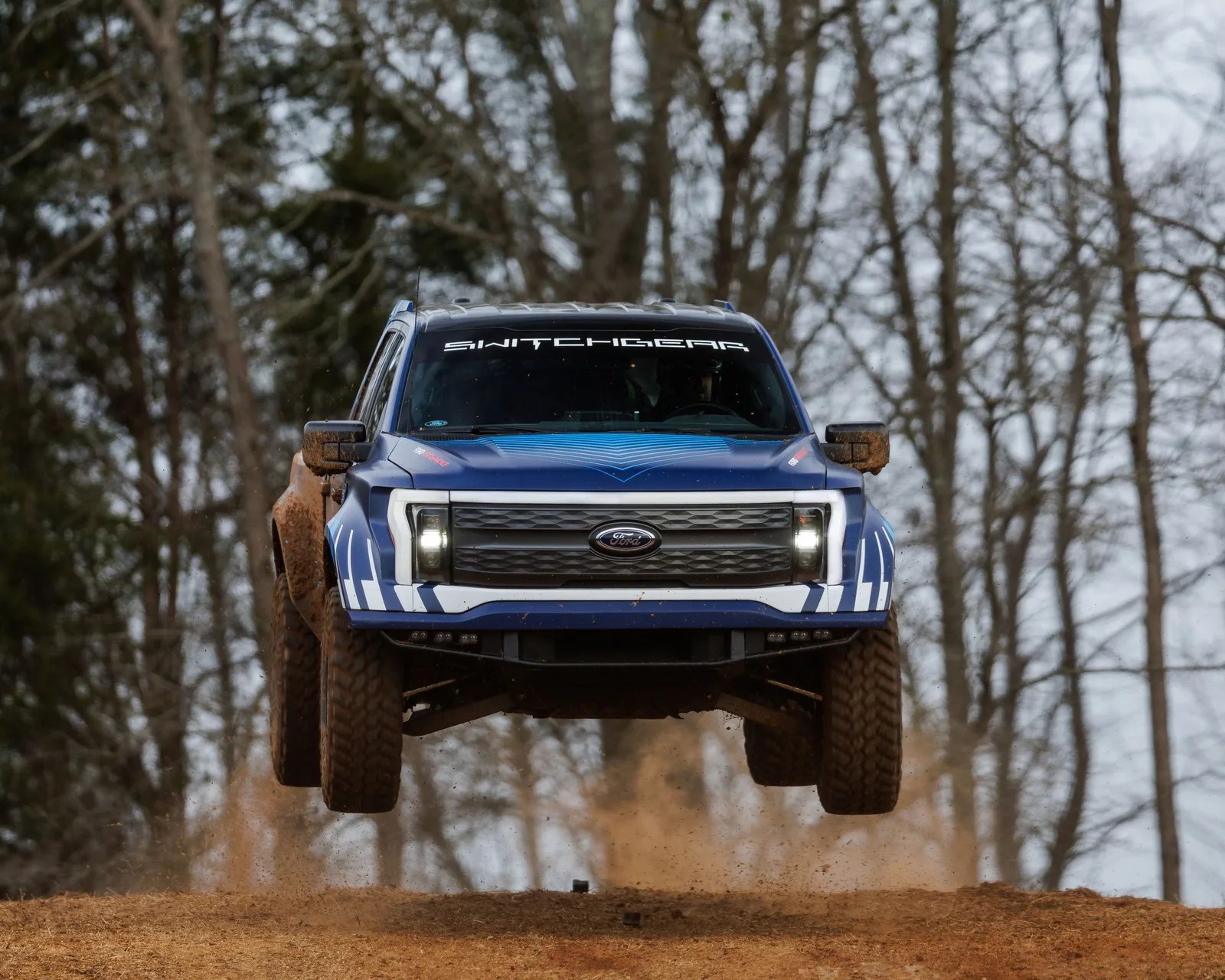 Blue Ford off-road truck with "SWITCHGEAR" decal on windshield airborne on dirt trail.