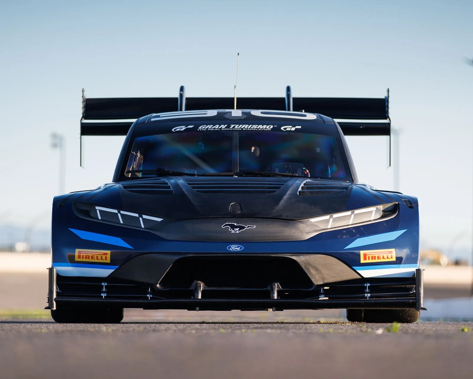 Front view of a blue and black Ford Mustang race car with Pirelli stickers and a large rear wing on a racetrack.