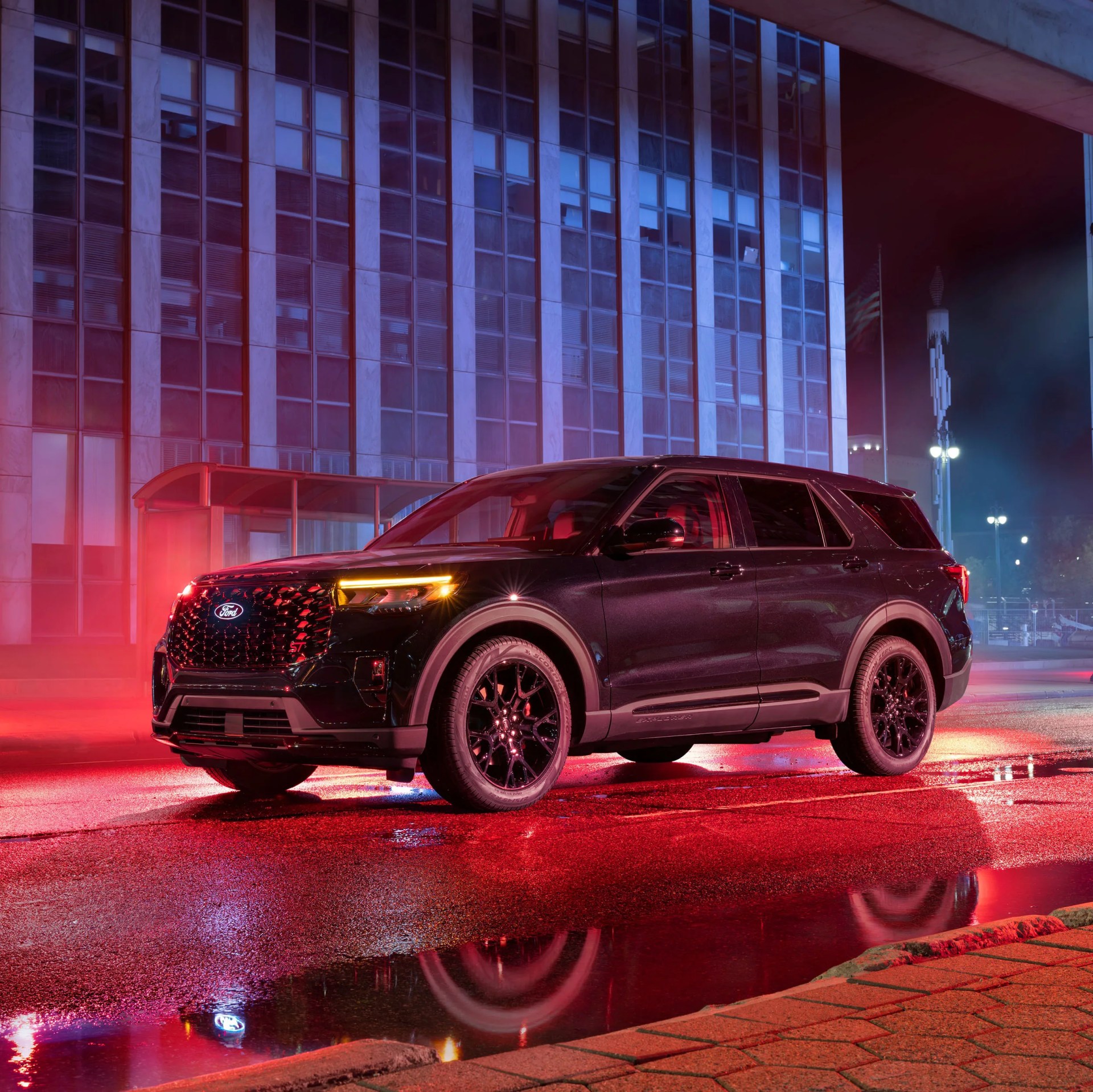 Black Ford SUV with illuminated headlights parked on a wet street at night in front of a tall building.
