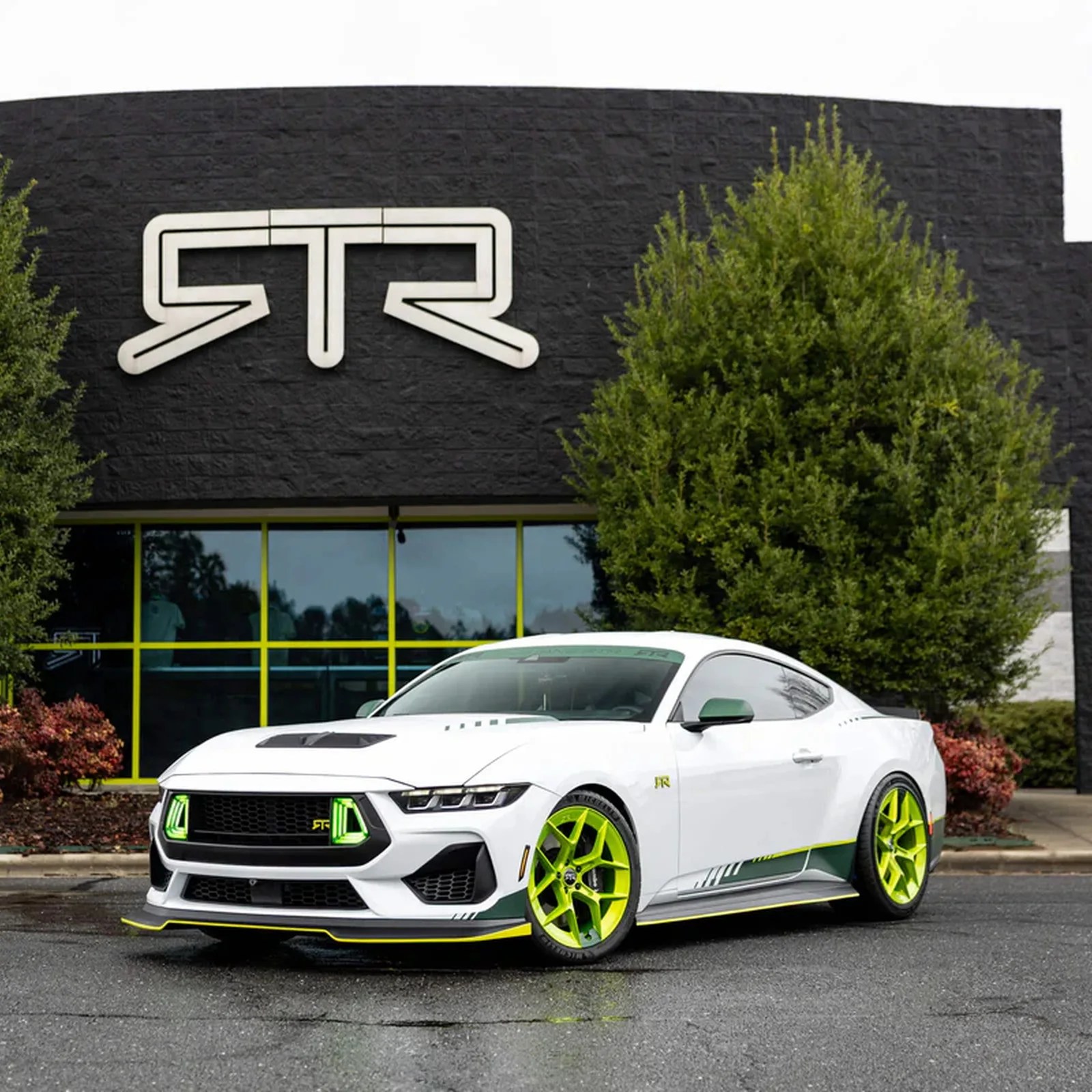 White sports car with neon green accents and wheels parked in front of a black building with a large "RTR" logo.