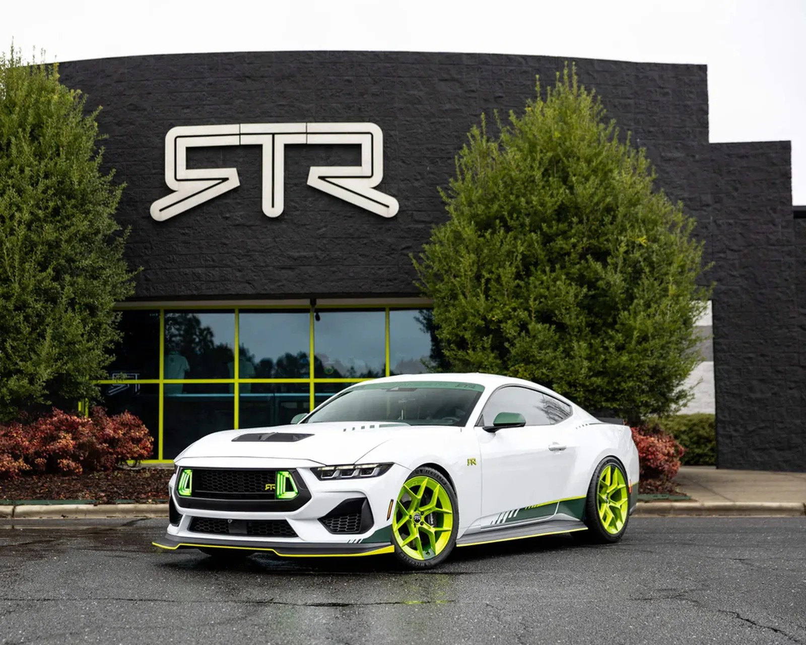 White sports car with neon green accents and wheels parked in front of a black building with a large "RTR" logo.