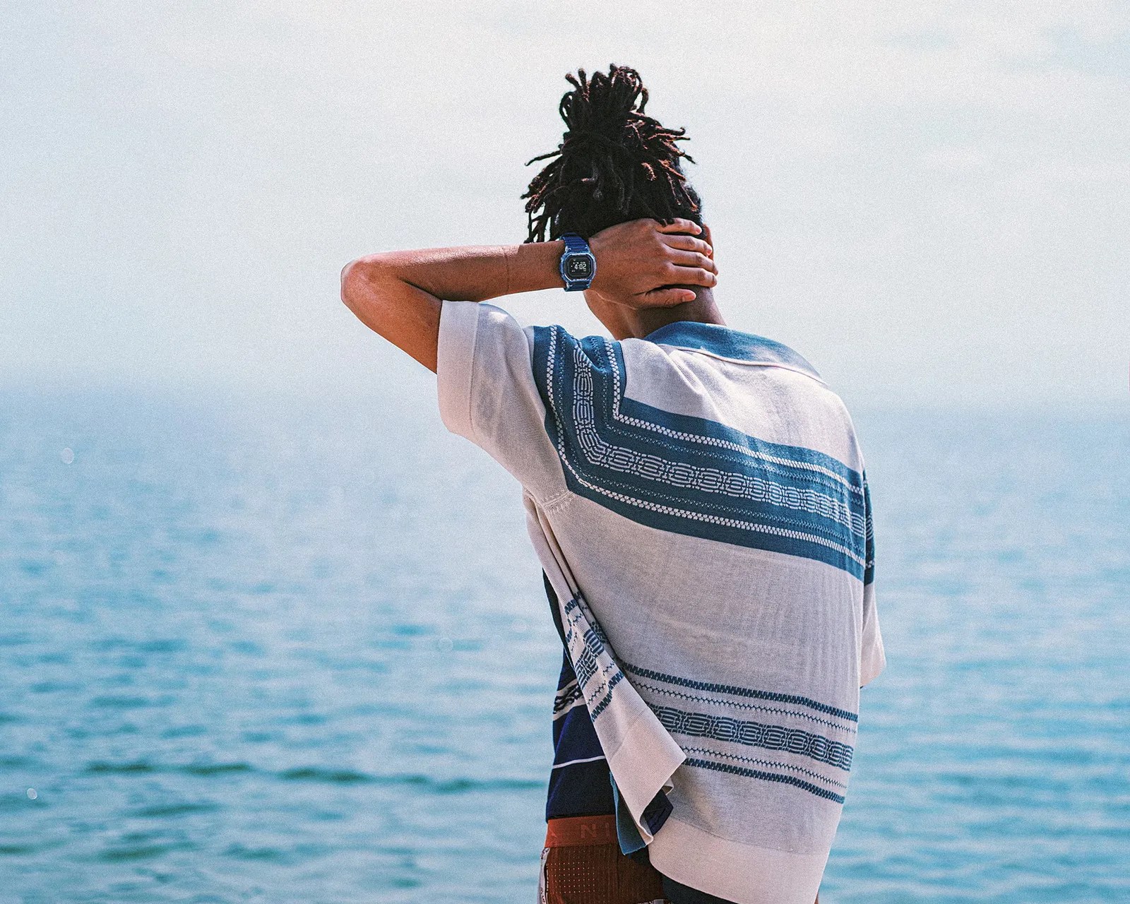 Person with dreadlocks wearing a blue and white patterned shirt and a digital watch, standing by the sea.