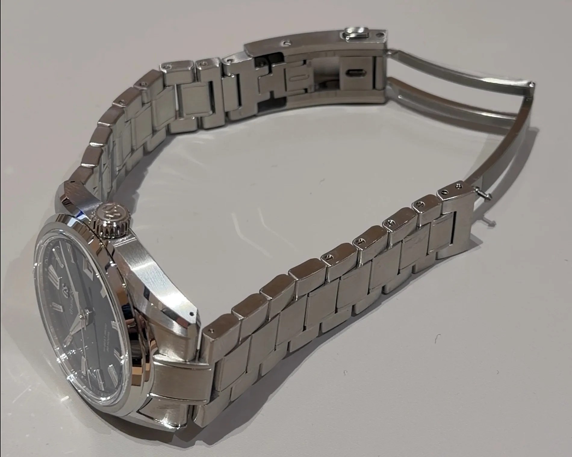 Silver metal watch band with a deployment clasp partially open, attached to a watch with a black dial.