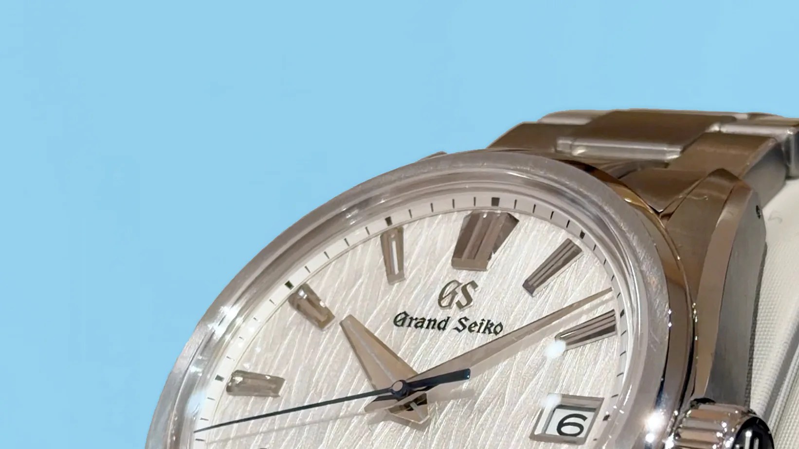 Close-up of a silver Grand Seiko wristwatch with textured dial, date window, and metal bracelet against a blue background.
