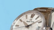Close-up of a silver Grand Seiko wristwatch with textured dial, date window, and metal bracelet against a blue background.