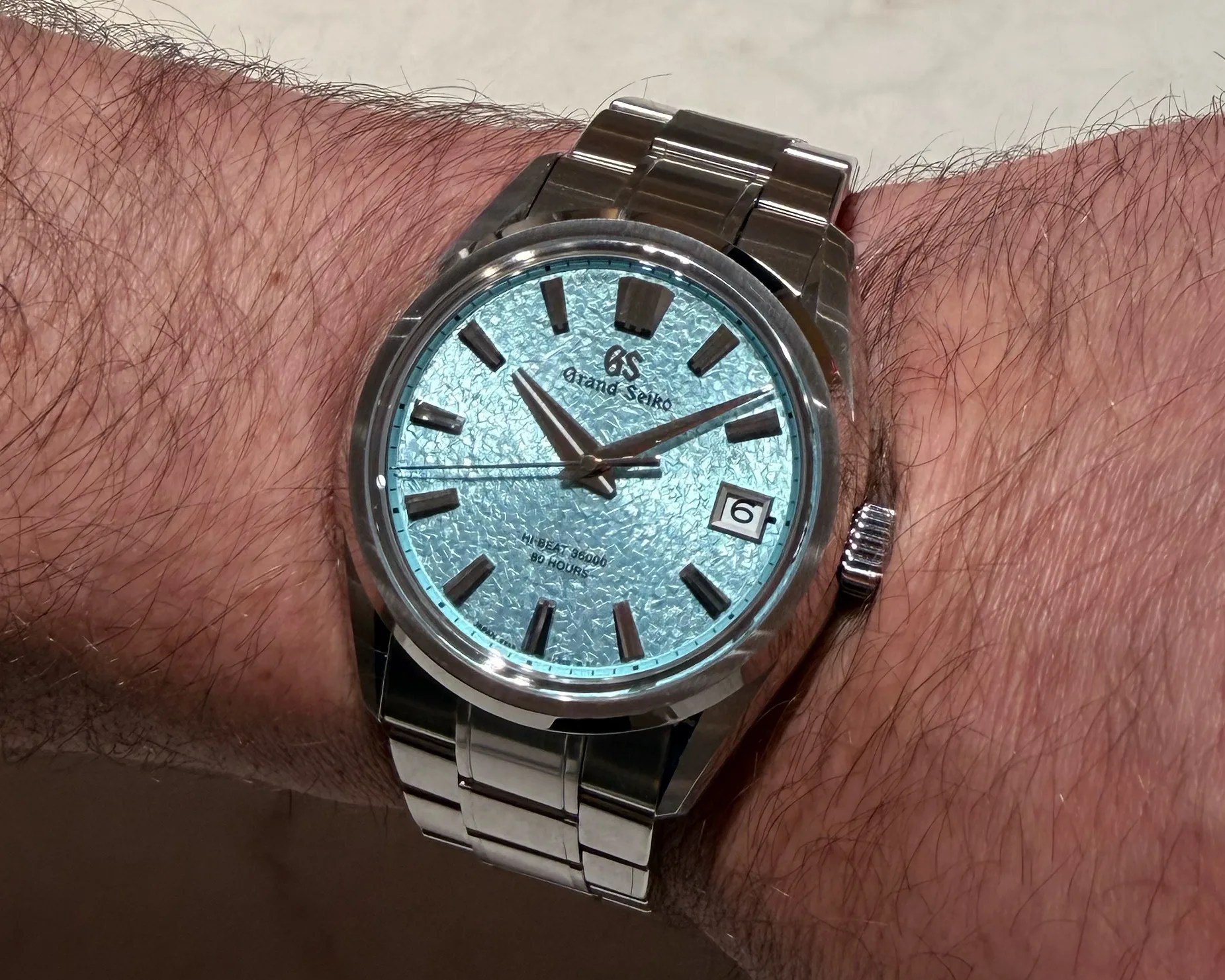 Grand Seiko wristwatch with textured light blue dial, silver hour markers, and date window on a metal bracelet.