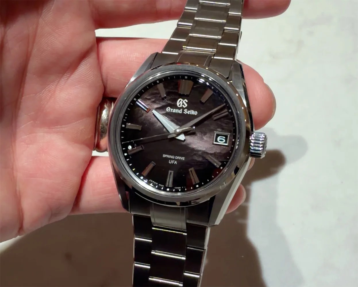 Grand Seiko wristwatch with textured dark dial, silver hour markers, date window, and stainless steel bracelet held in a hand.