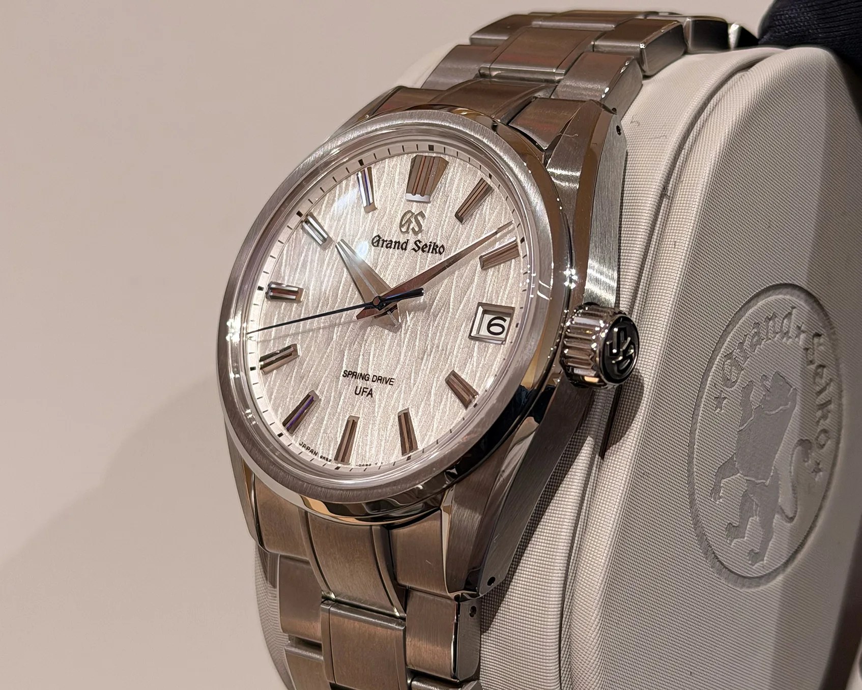 Silver Grand Seiko wristwatch with textured dial, date window, and metal bracelet on a white cushion.
