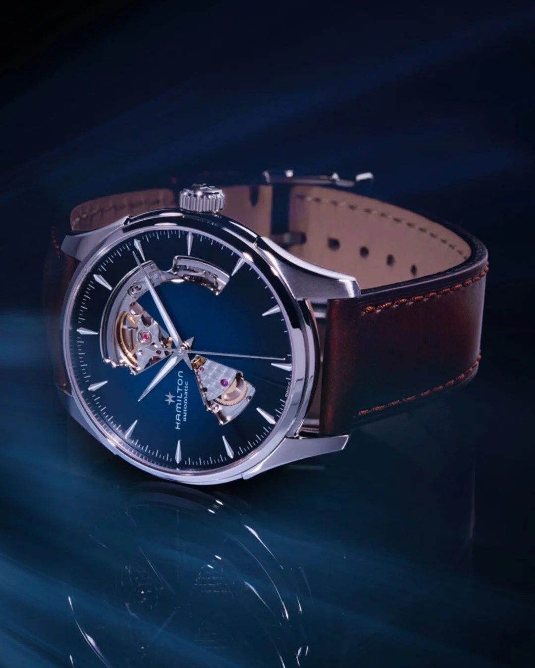 Hamilton automatic wristwatch with blue dial, silver case, and brown leather strap on a dark reflective surface.