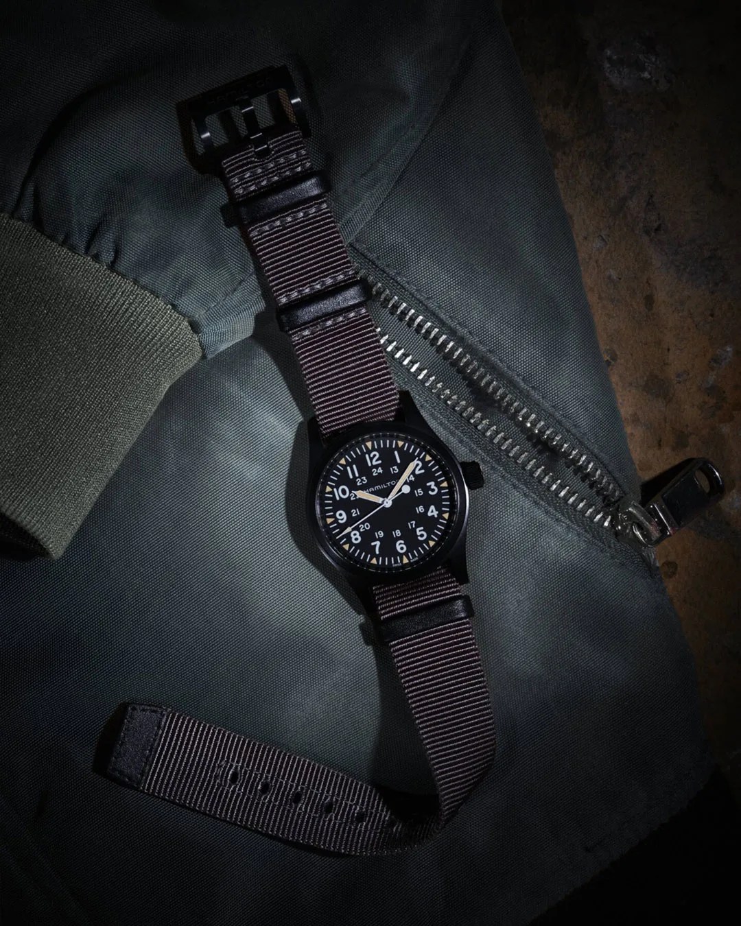Black Hamilton wristwatch with white numerals and a dark brown nylon strap placed on a green jacket with a silver zipper.
