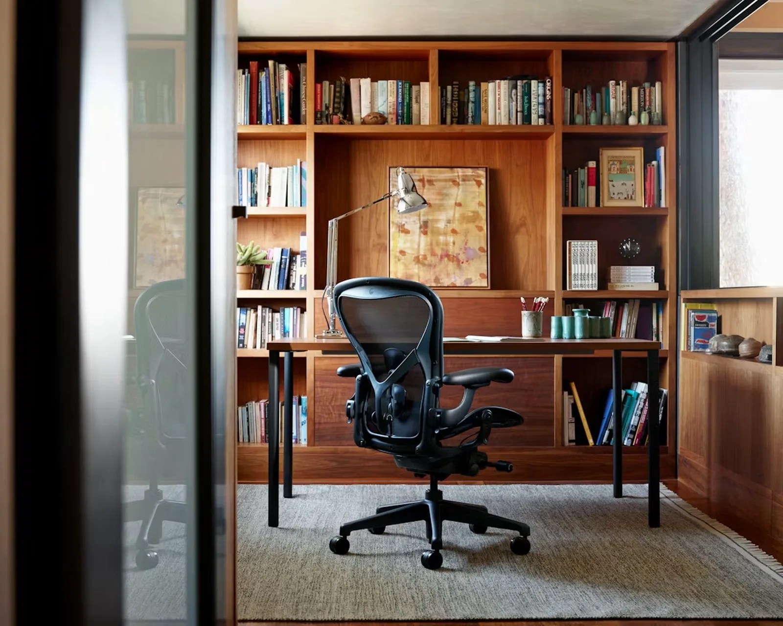 Black ergonomic office chair in front of a wooden desk with a metal lamp and built-in wooden bookshelves filled with books.