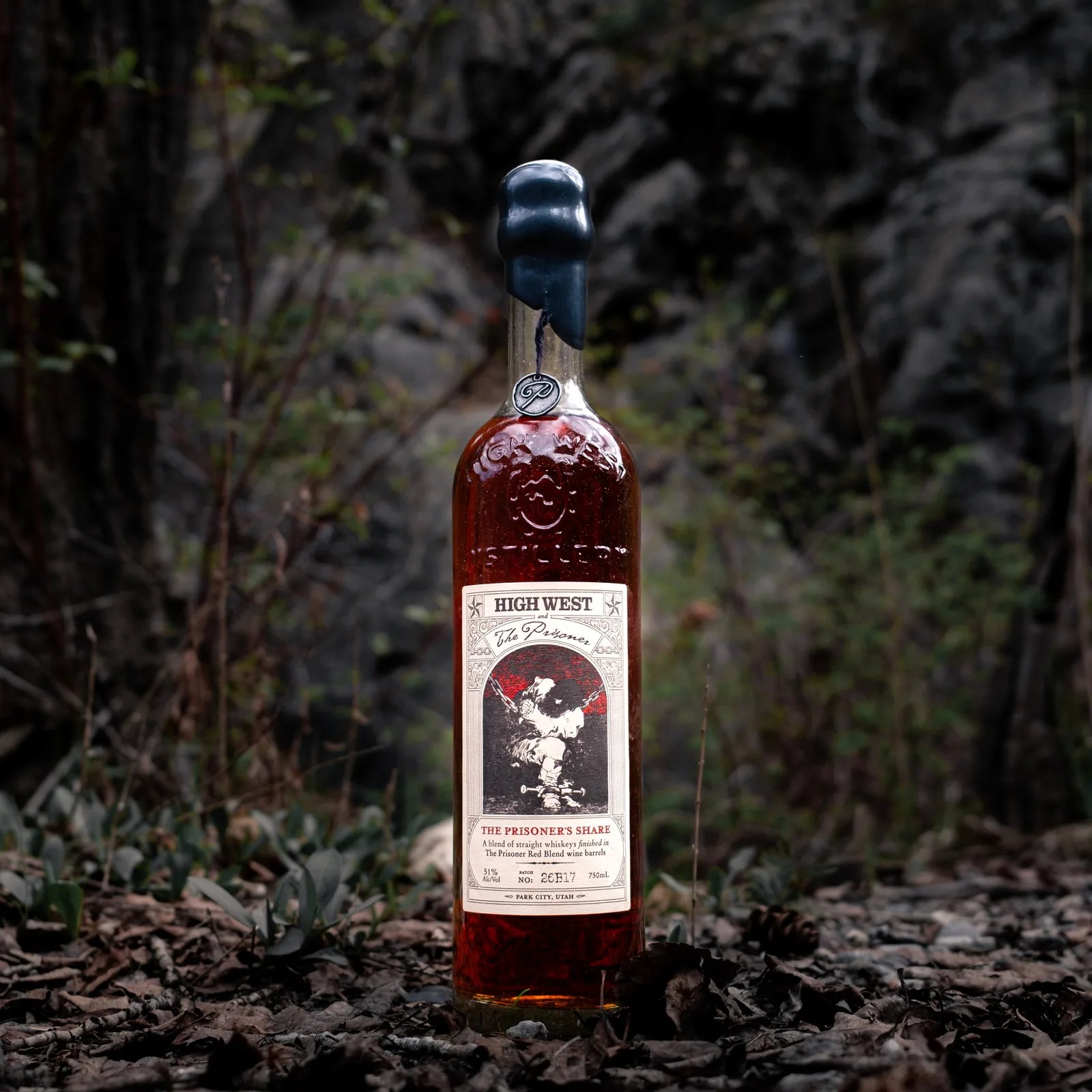 Tall glass bottle of High West whiskey with black wax seal on a forest floor with leaves and rocks.