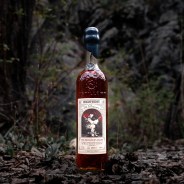 Tall glass bottle of High West whiskey with black wax seal on a forest floor with leaves and rocks.
