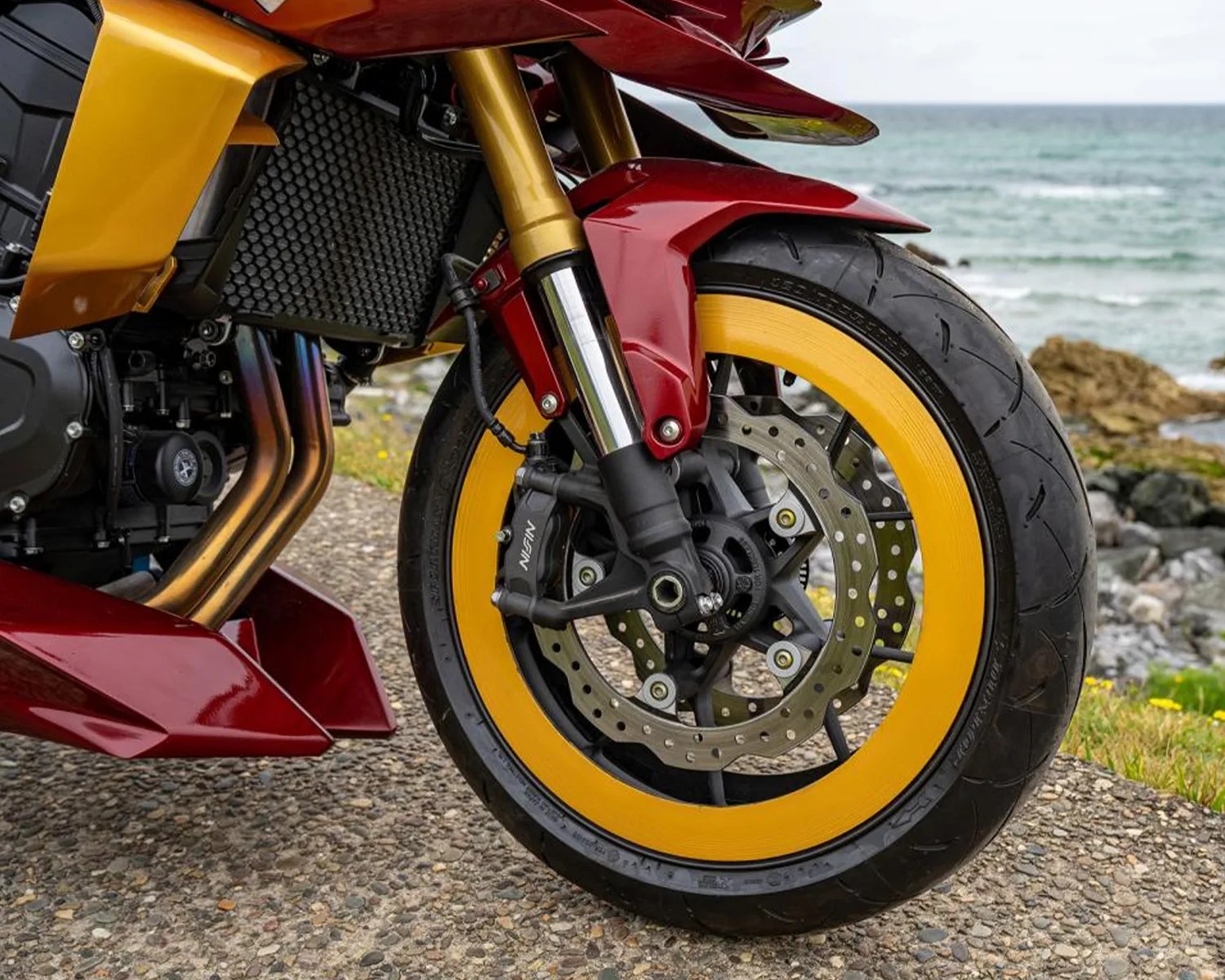Front wheel of a motorcycle with yellow rim tape, red fender, and gold suspension forks on a rocky path by the sea.
