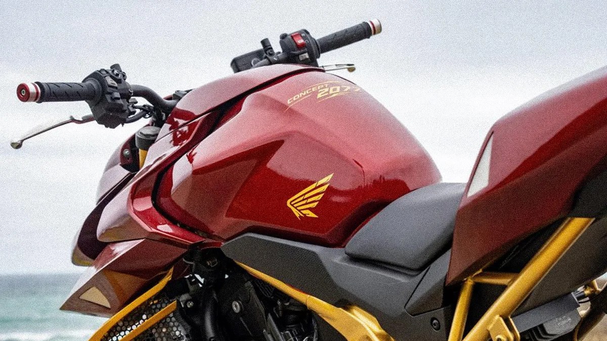 Close-up of a red and gold Honda motorcycle fuel tank and seat with black handlebars against a cloudy sky.