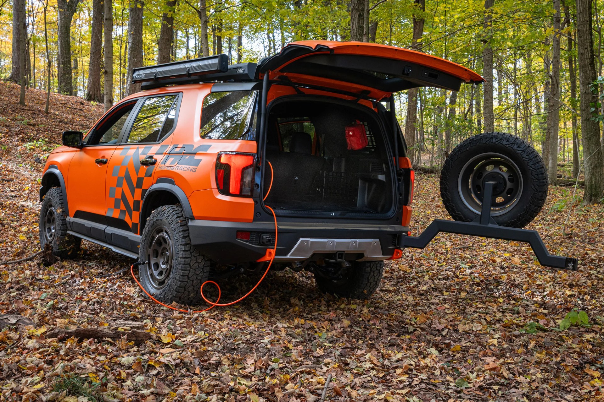 Orange off-road SUV with rear hatch open, spare tire mounted on swing arm, and orange cable plugged in, parked in a forest.