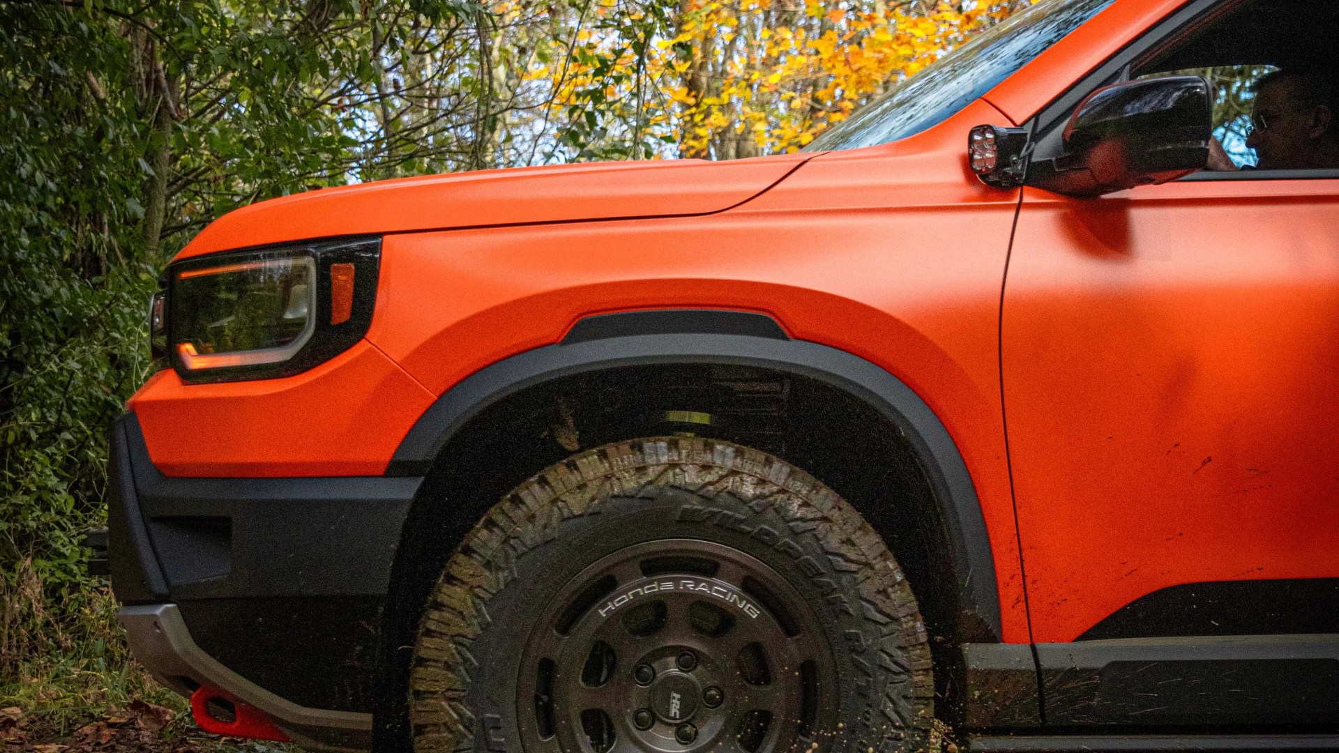 Orange off-road vehicle front side with large mud tires and black fender flares in a wooded area.