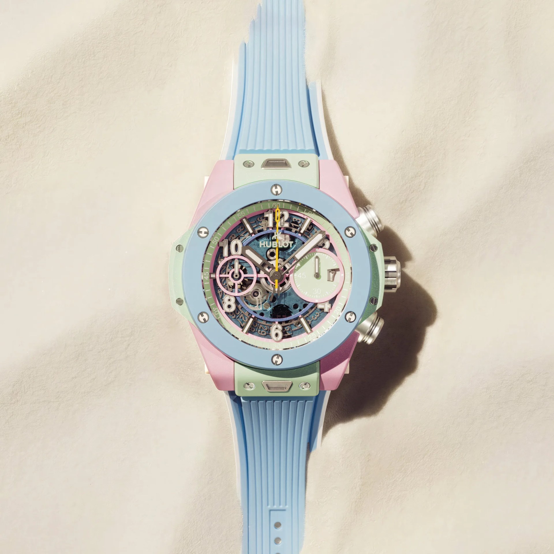 Pastel-colored Hublot wristwatch with blue rubber strap and visible mechanical gears on a beige surface.