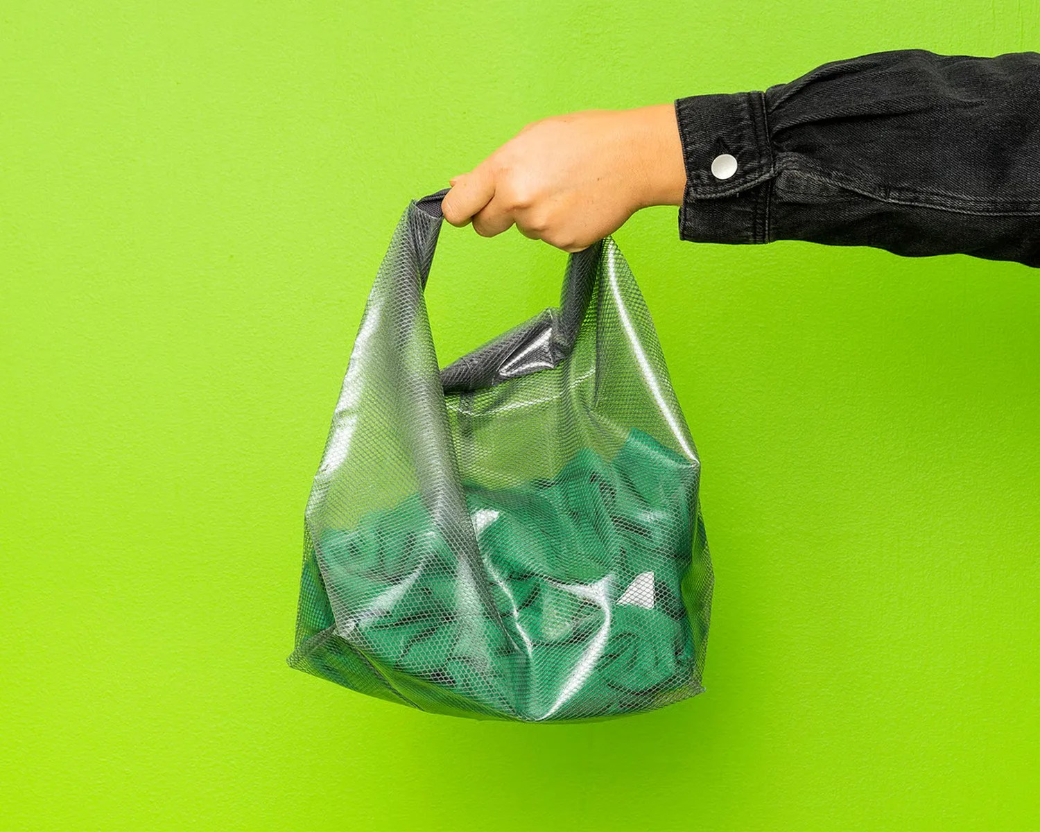 Hand holding a shiny silver mesh bag containing green fabric against a bright green wall.