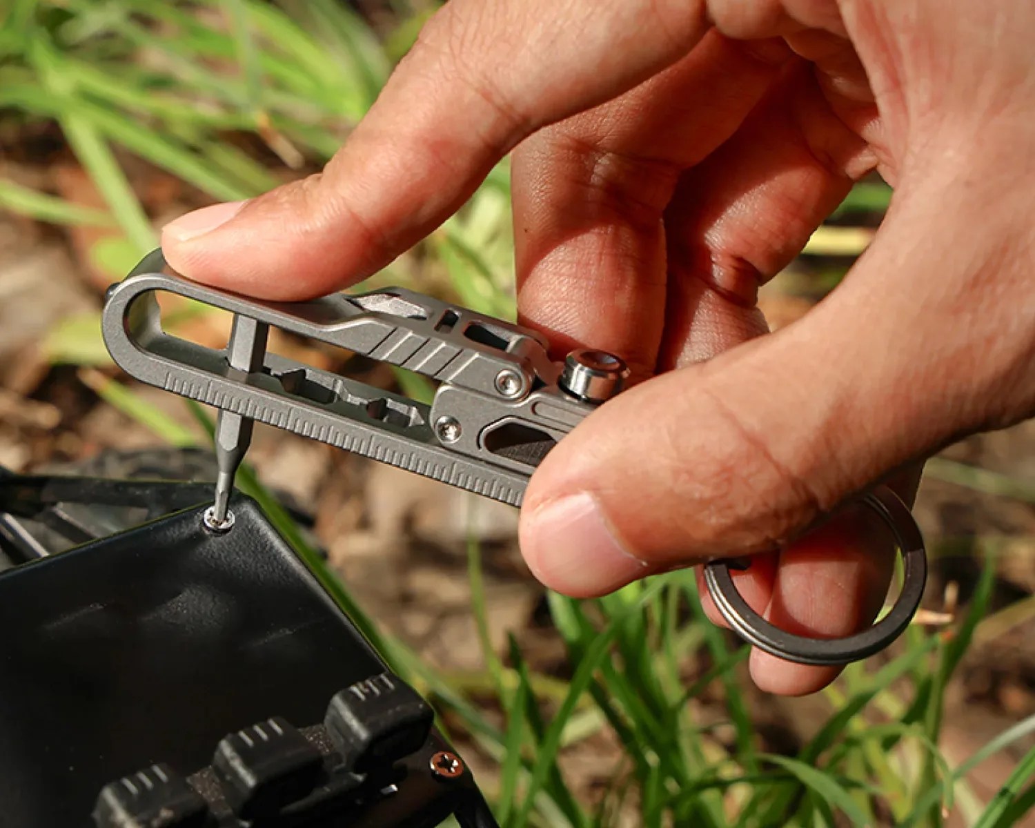 Hand holding a compact metal multi-tool with a screwdriver bit tightening a screw on a black device outdoors.