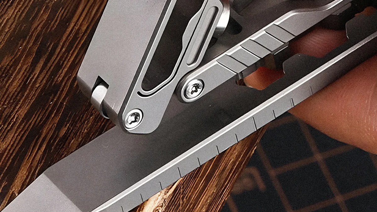 Close-up of a silver folding knife blade with measurement markings and a metal clip held in a hand.