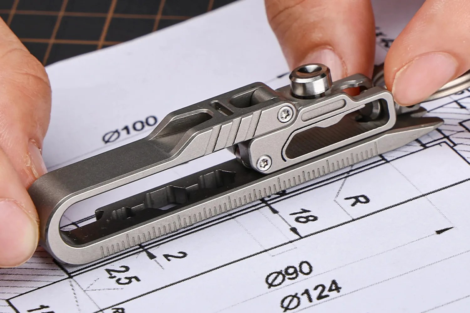 Compact titanium multi-tool with ruler and various attachments held over a technical drawing.