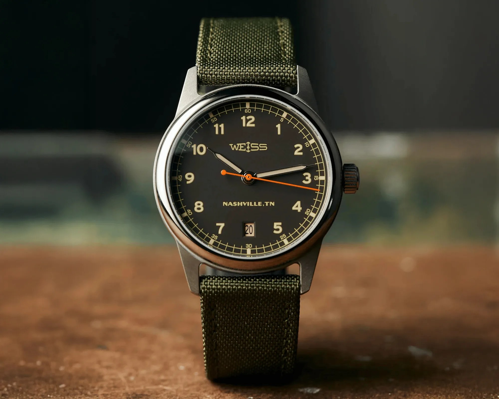 Black Weiss wristwatch with beige numerals, orange second hand, date window, and green fabric strap.
