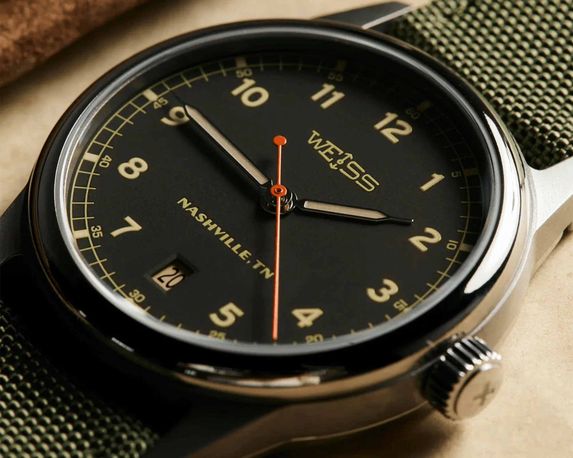 Close-up of a black Weiss wristwatch with beige numerals, orange second hand, and green fabric strap.