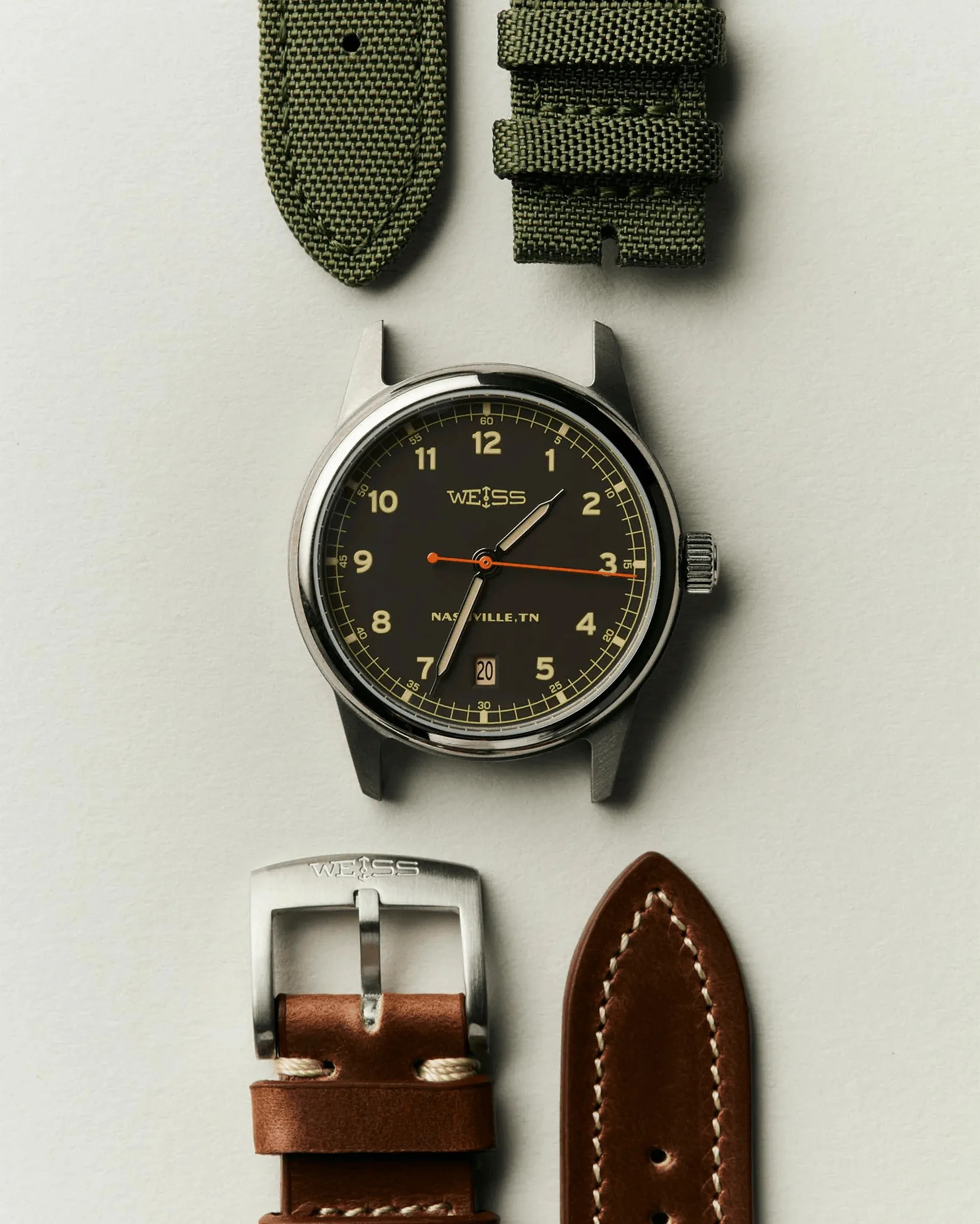 Watch face with black dial and beige numerals, two green fabric strap pieces above, and brown leather strap pieces with silver buckle below.
