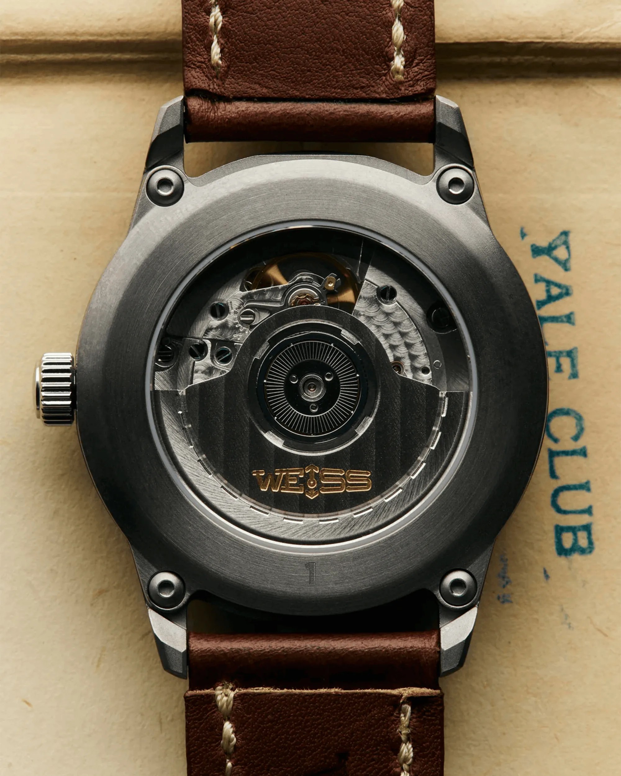 Close-up of the back of a Weiss watch with visible mechanical movement and brown leather strap.