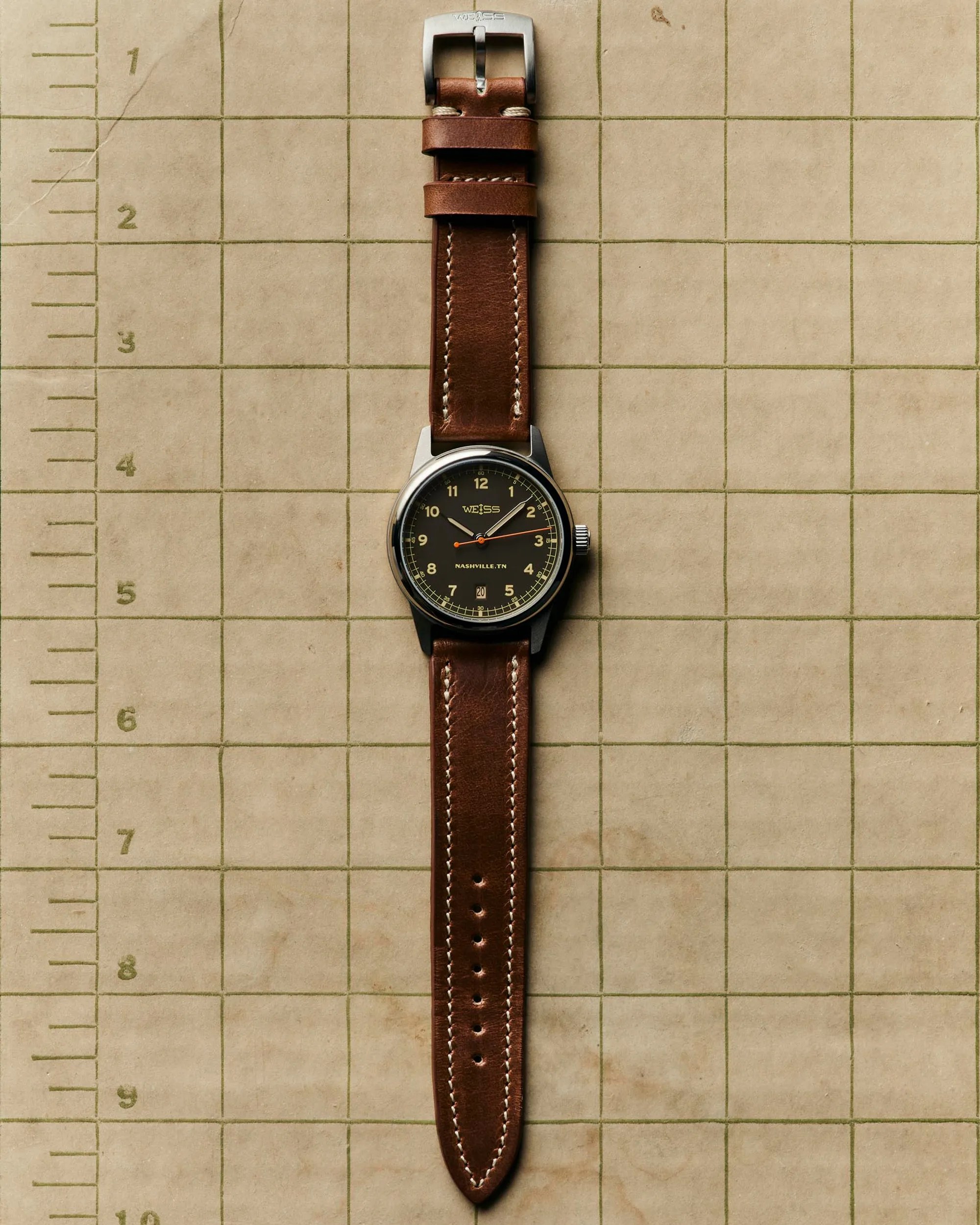 Brown leather watch strap with white stitching and a black Weiss watch face on a grid-patterned beige background.