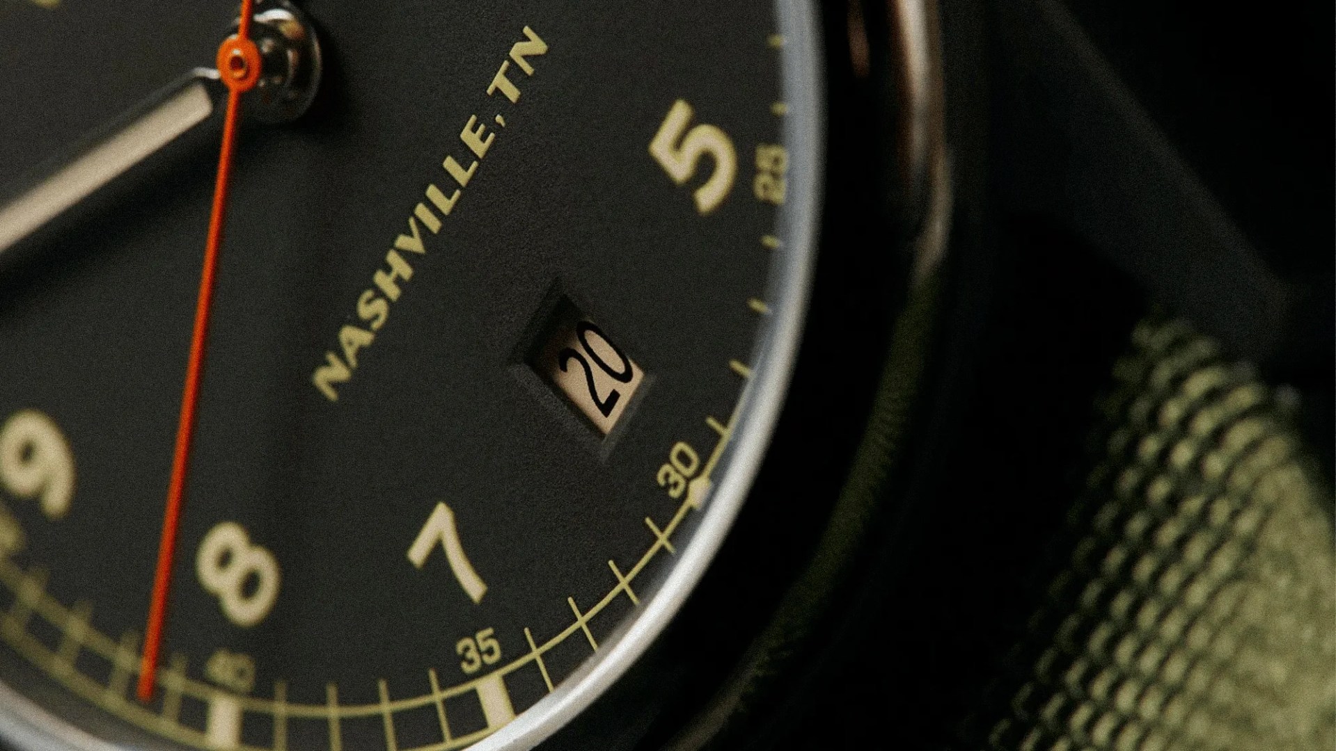 Close-up of a black watch face showing the date "20," with beige numerals and an orange second hand.