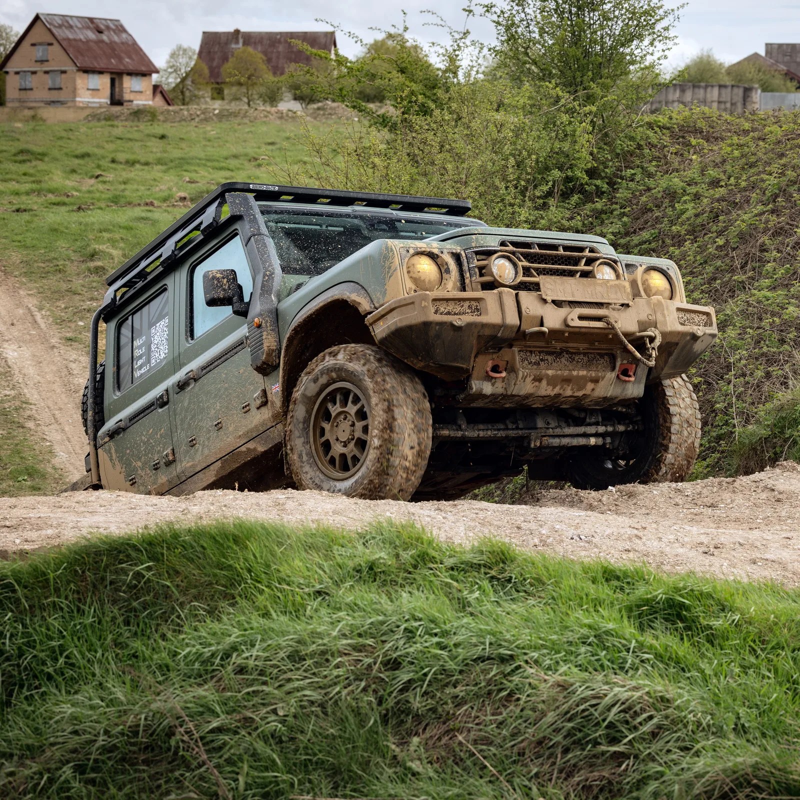 Green off-road vehicle with mud splatters driving on a dirt path near grassy hills and houses.