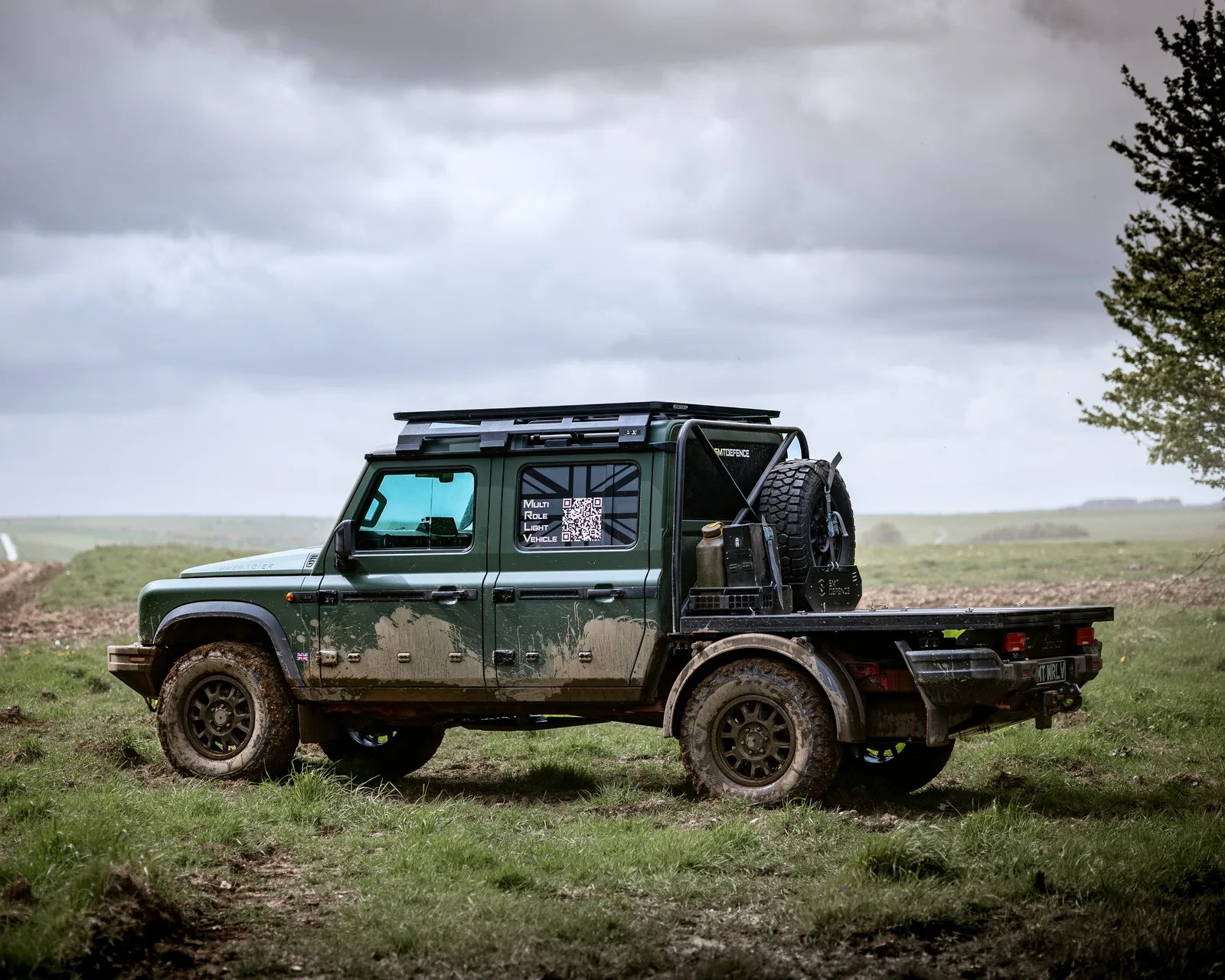 Green off-road pickup truck with mud splatters and spare tire mounted on the flatbed, parked on grass under cloudy sky.