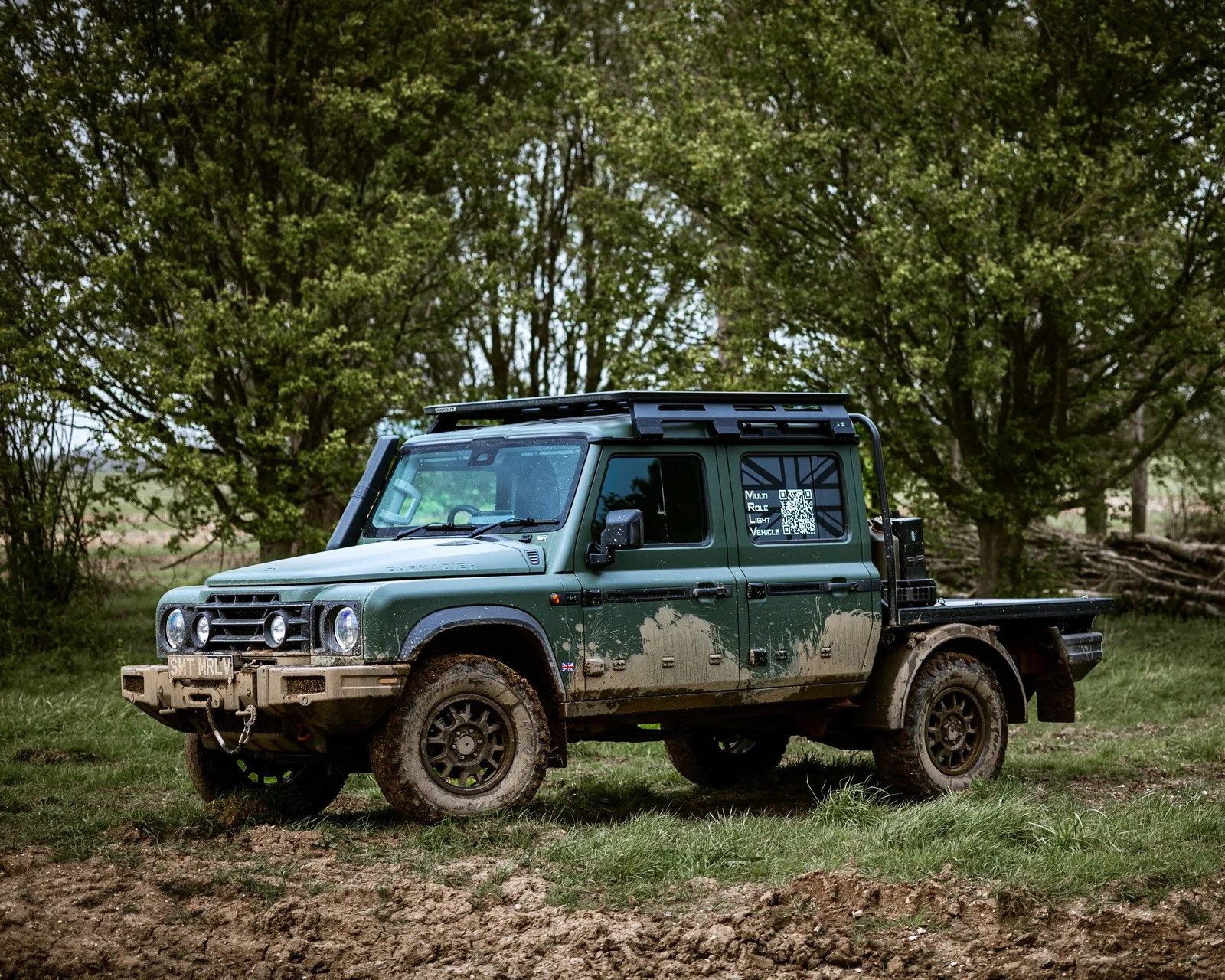 Green off-road pickup truck with mud splatters parked on grassy terrain with trees in the background.