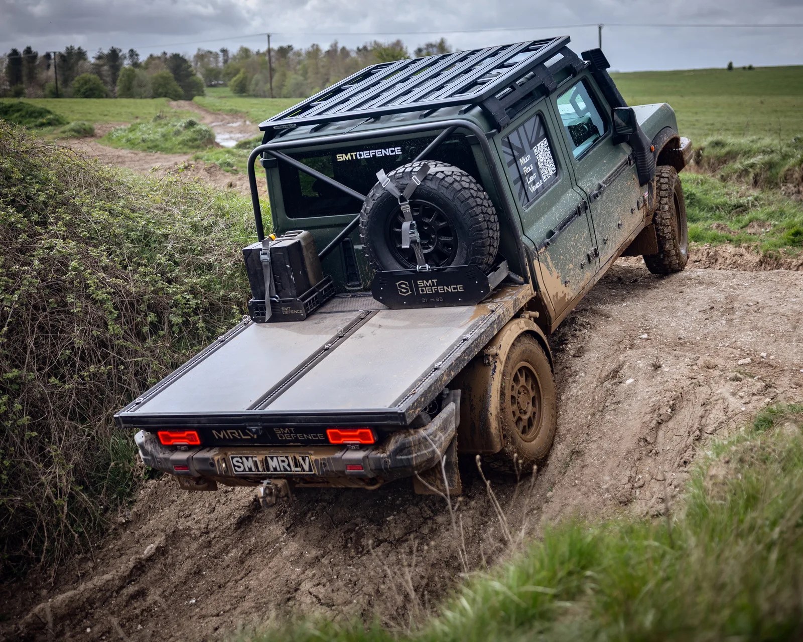 Green off-road pickup truck with flatbed, spare tire, and equipment boxes driving on muddy terrain.