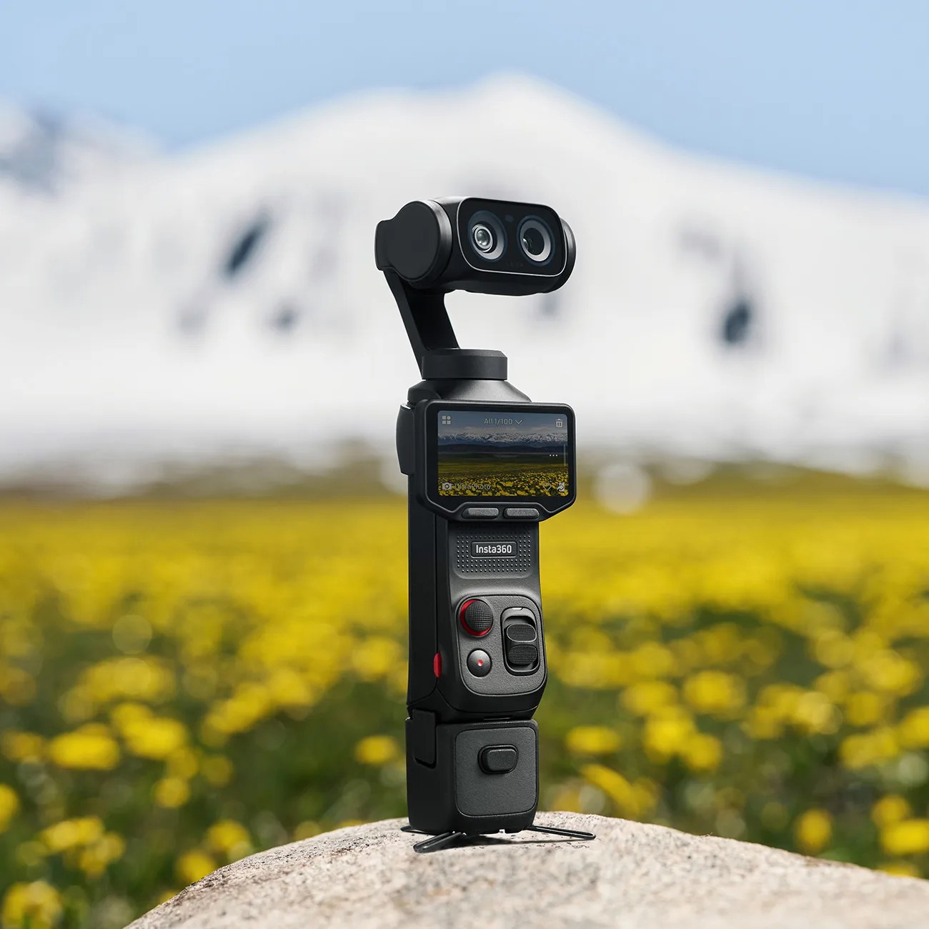 Black Insta360 camera with dual lenses and screen, standing on a rock in a yellow flower field with snowy mountains in the background.