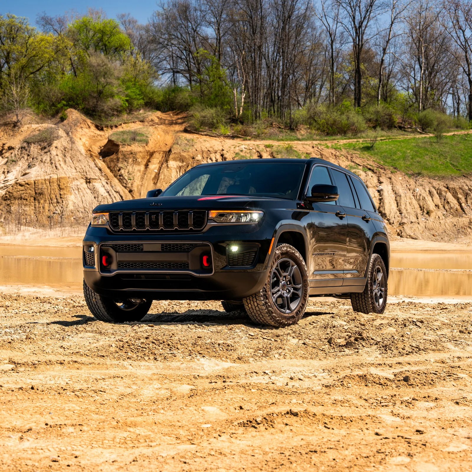 Black Jeep SUV with red tow hooks parked on sandy terrain with trees and a dirt hill in the background.