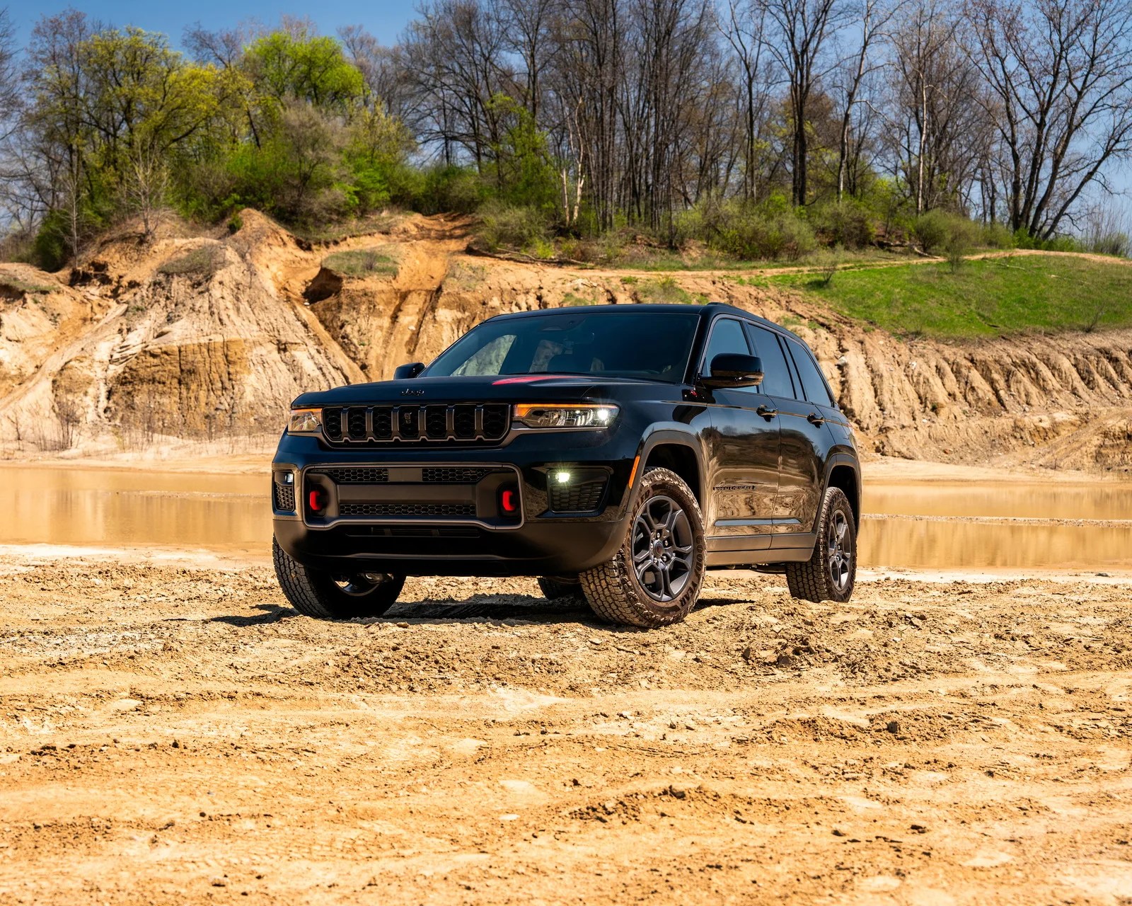 Black Jeep SUV parked on dry, sandy terrain with muddy water and trees in the background.