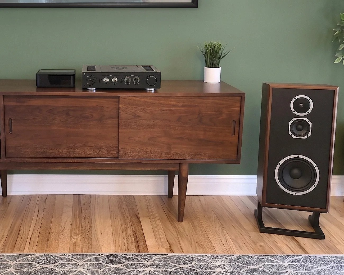 Mid-century wooden sideboard with black audio equipment and a potted plant on top, next to a large wooden speaker on a stand.