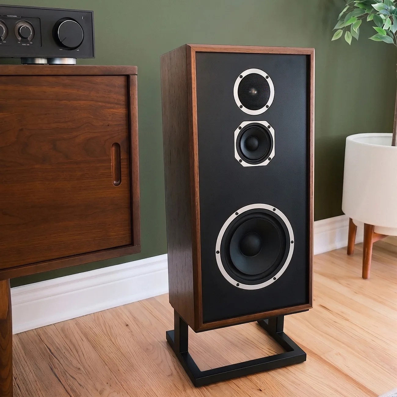 Wooden floor-standing speaker with black front panel and three drivers on a black metal stand next to a wooden cabinet.