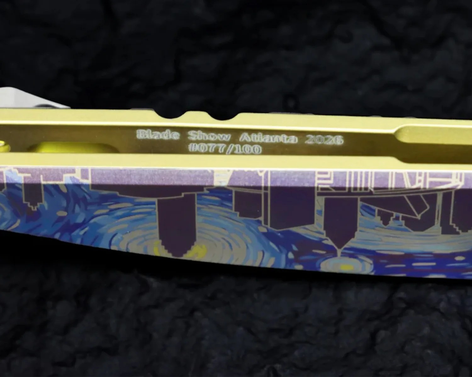 Close-up of a gold-colored knife spine engraved with "Blade Show Atlanta 2026 #077/100" and a colorful cityscape design on the handle.