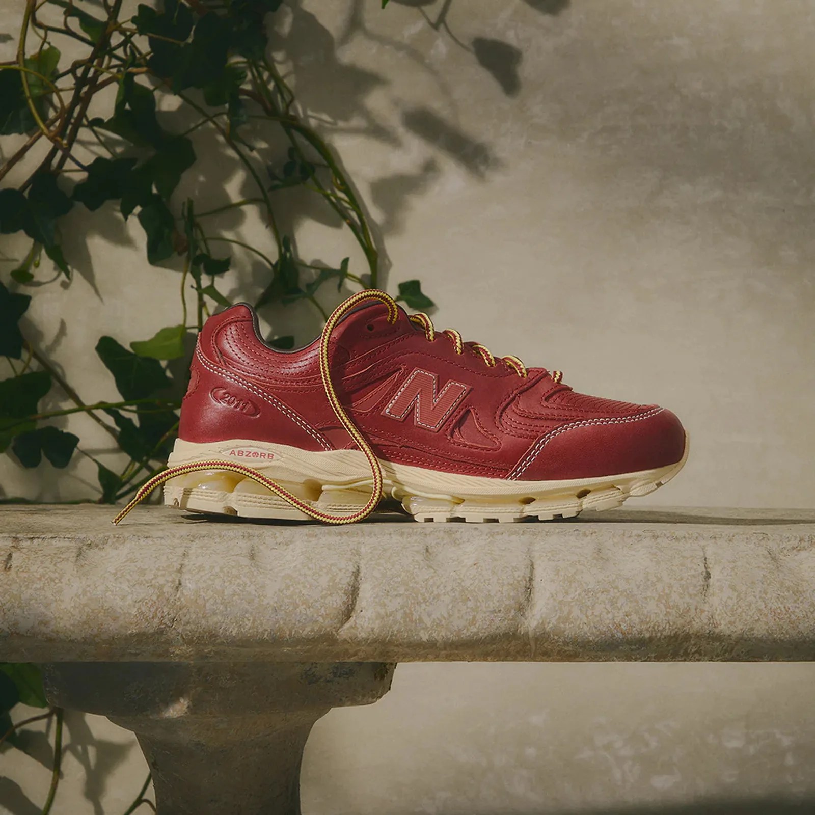 Red New Balance sneaker with yellow and red laces on a stone ledge with ivy shadows in the background.
