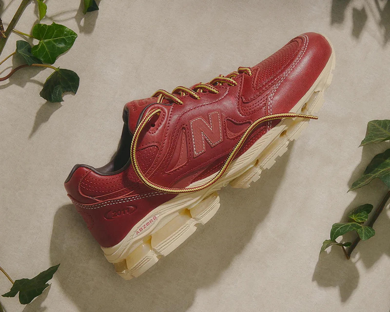Red New Balance sneaker with yellow and red laces and beige sole on a light surface with green leaves.