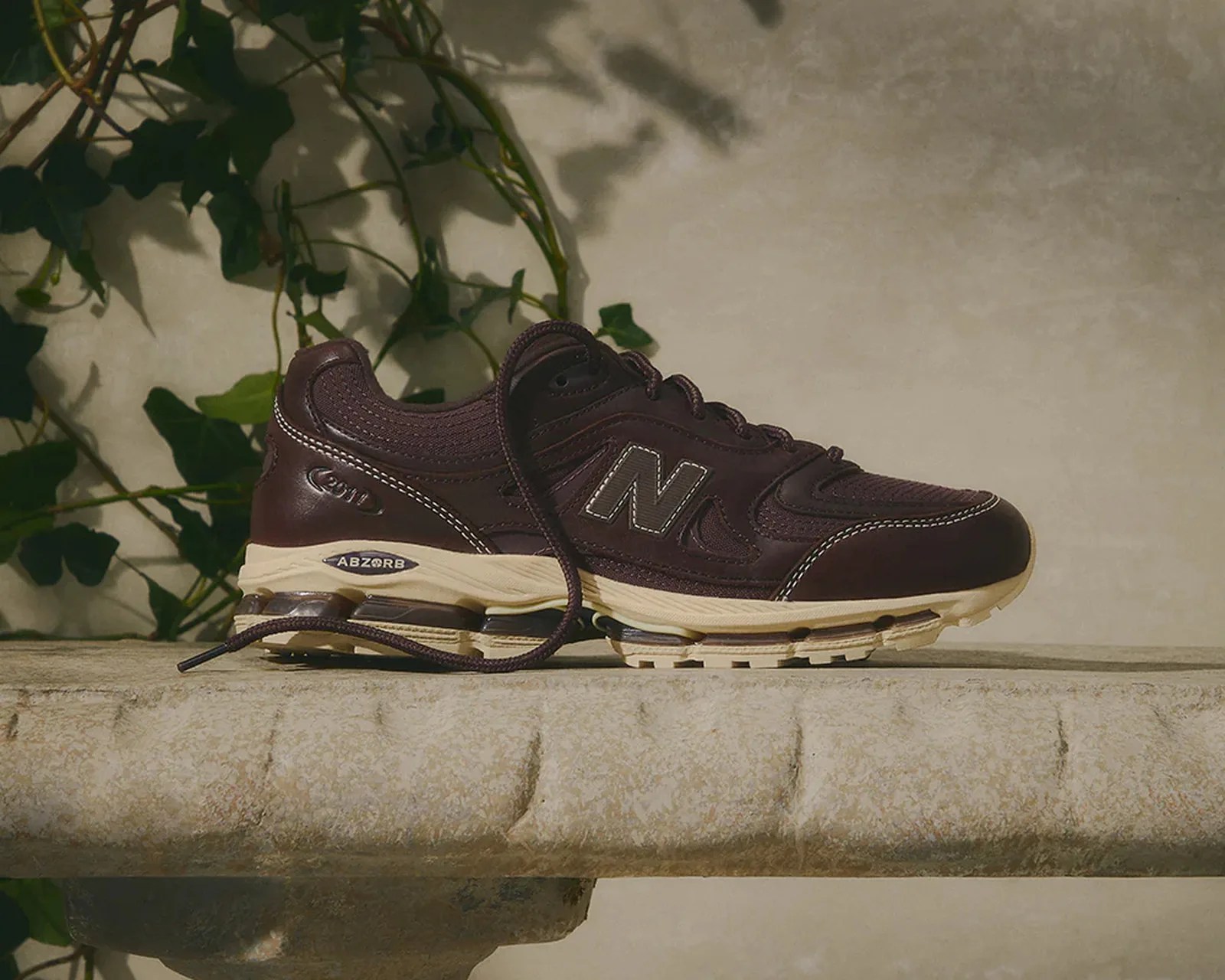 Dark brown New Balance sneaker with beige sole and visible cushioning, placed on a stone surface with green leaves in the background.