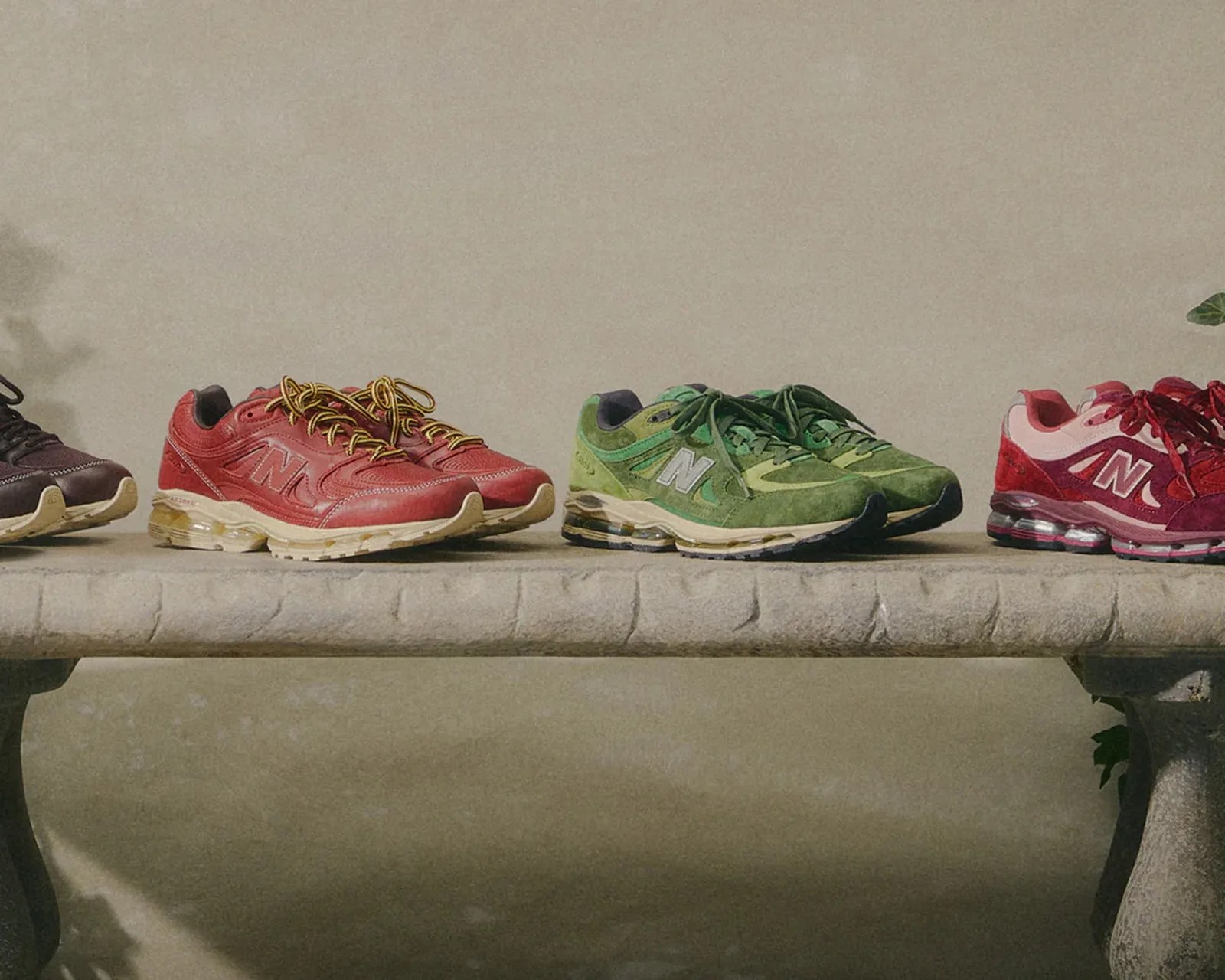 Four pairs of New Balance sneakers in dark brown, red, green, and red with pink accents, displayed on a stone bench.