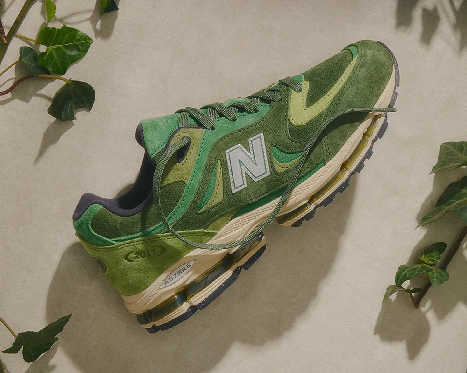 Green New Balance sneaker with suede and mesh panels, white midsole, and visible cushioning on a beige surface with green leaves.