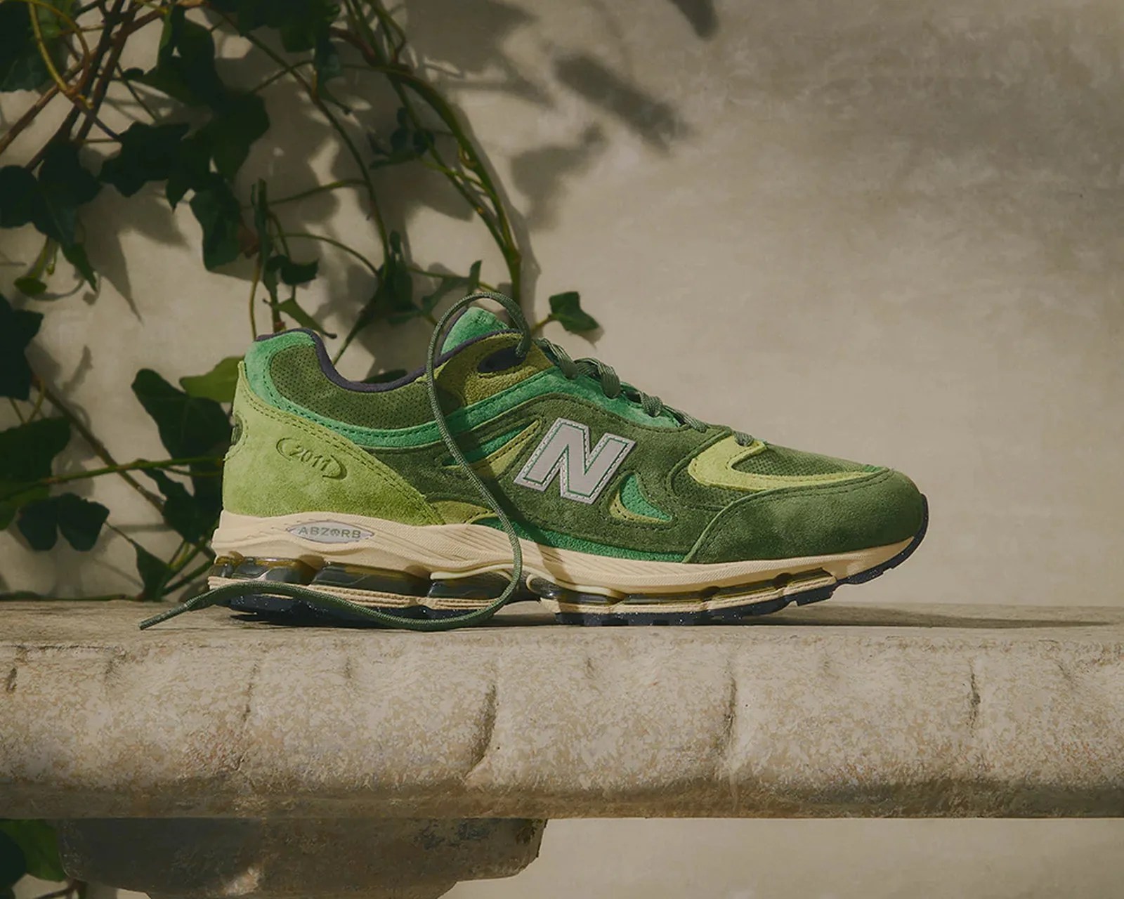 Green suede New Balance sneaker with beige sole and visible cushioning on a stone ledge.