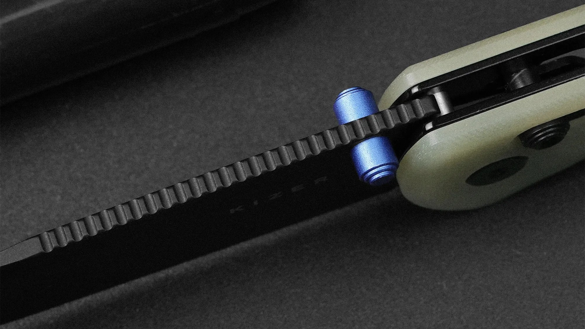 Close-up of a black folding knife with a textured blade spine and a blue thumb stud on a dark surface.
