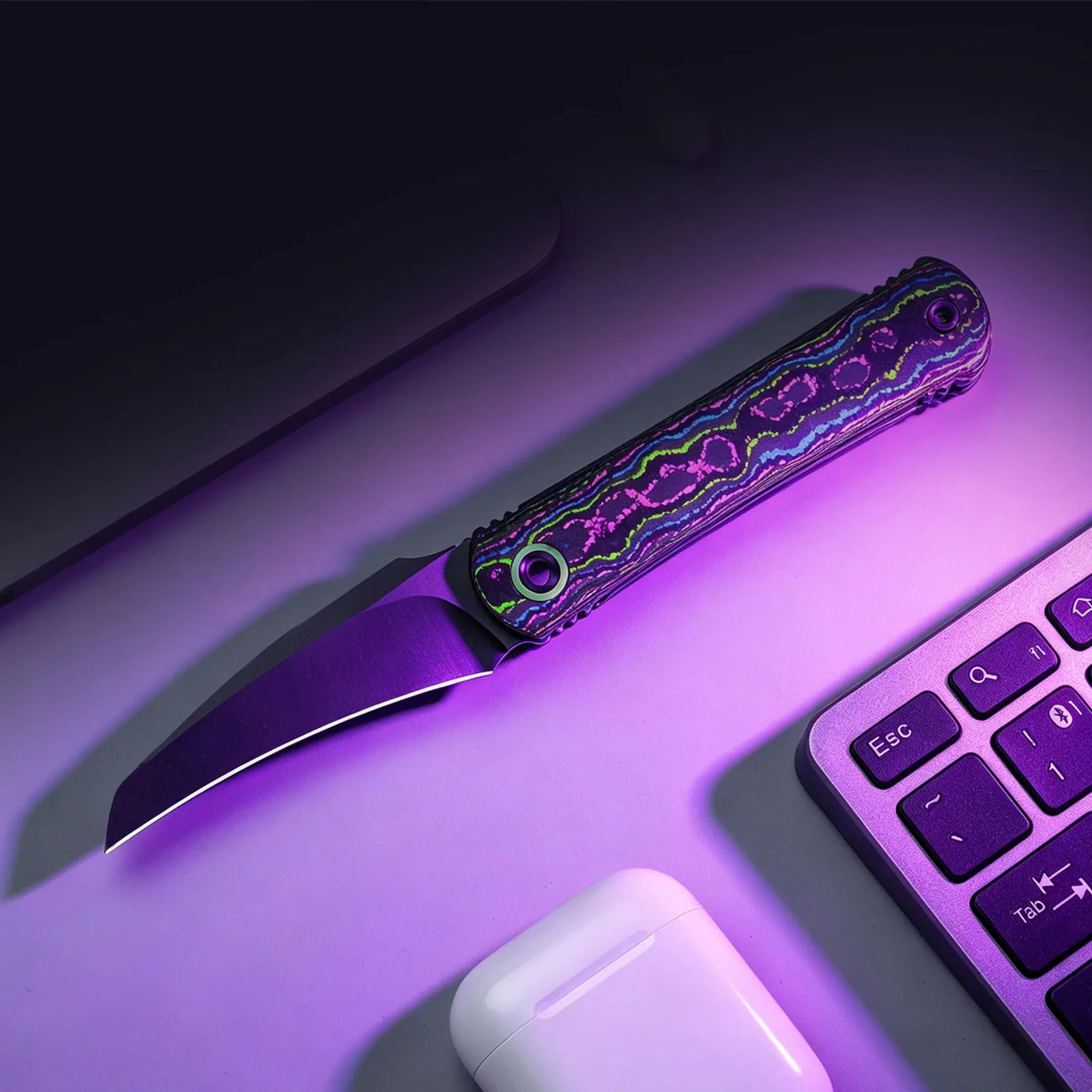 Folding knife with a colorful patterned handle next to a laptop keyboard and white wireless earbud case under purple light.